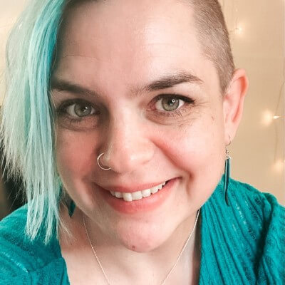 Joy is a white woman. Her hair is shaved on her left side and long and turquoise on the right. She's wearing a turquoise top and smiling at the camera.